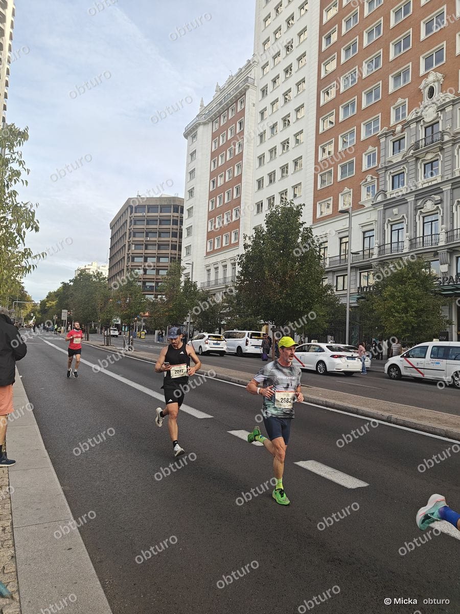 Plusieurs participants courent sur une route urbaine bordée de bâtiments sous un ciel légèrement nuageux.