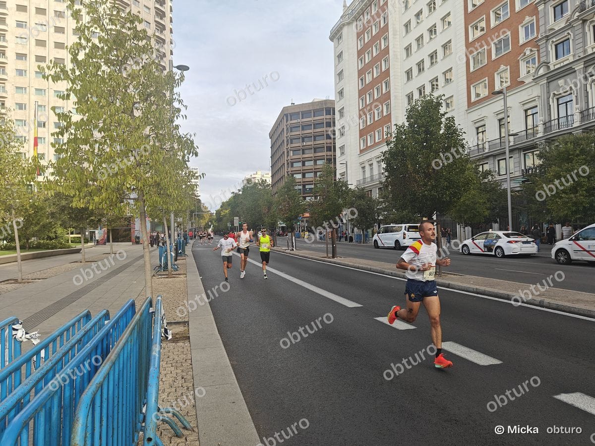 Plusieurs participants courent sur une route urbaine réservée, entourés de bâtiments et d'arbres, sous un ciel légèrement nuageux.